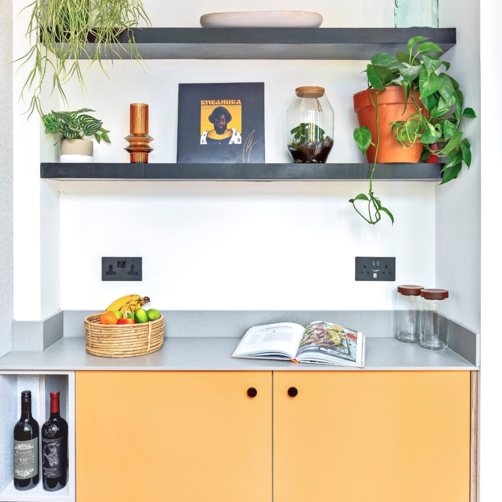 kitchen corner with orange cabinets and grey marble worktop, black sockets and shelving with plants and art