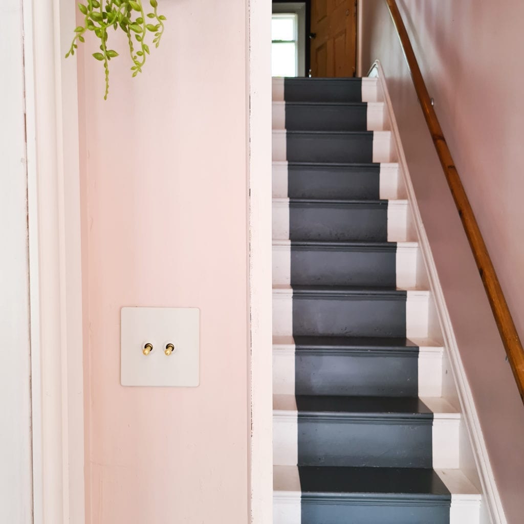white and gold double toggle switch on soft pink wall in a hallway besides a flight of dark grey and white painted wooden stairs
