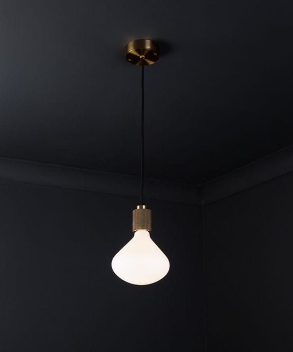 gold lanesborough pendant ceiling light suspended from a gold ceiling rose with black fabric cable and opal light bulb against a black wall