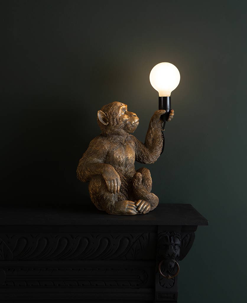 Koko gold sitting monkey table lamp on dark background holding switched on lightbulb