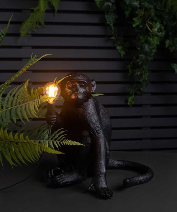 monty monkey light holding a lit bulb sat on top of a black table against black fencing