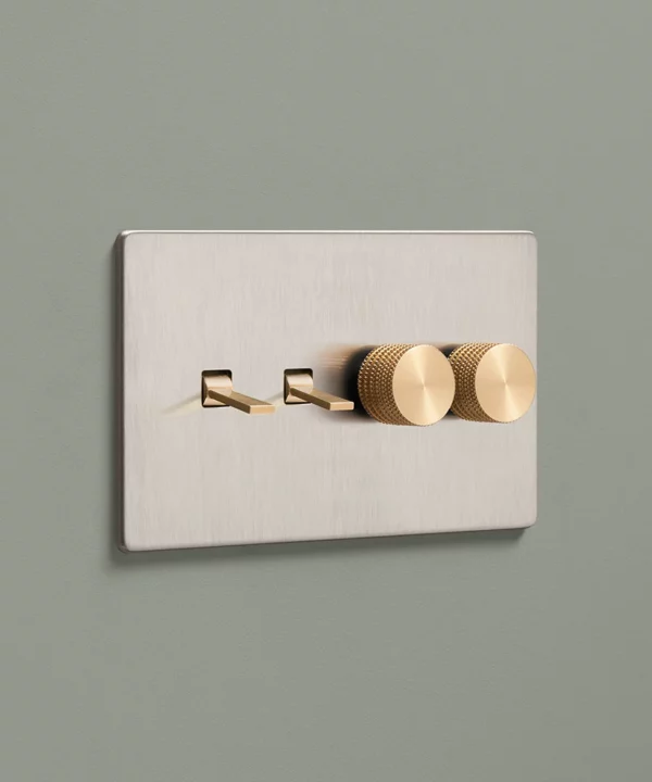 brushed silver combination light switch with two tapered toggles and two knurled dimmer knobs in gold mounted on a dusty sage painted wall