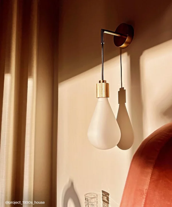A gold art deco inspired wall light with a frosted teardrop shaped bulb against a peach wall.